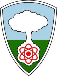 A shield with a white mushroom cloud rising from a red atom against a blue sky