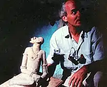 A balding white man, his face half obscured by shadow, poses next to a stone figurine.