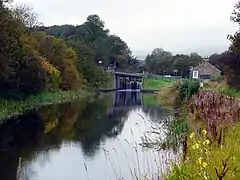 A view along a canal with a road bridge in the foreground and a canal lock beyond the bridge. Trees and other water plants align the canal.