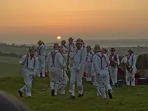 A group of twelve individuals wearing all-white costumes, including hats, are dancing in the centre of a green, grassy space. The low position of the sun indicates that it is early in the morning