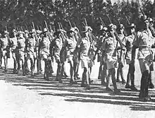 Soldiers in colonial-era military uniform march with rifles shouldered.