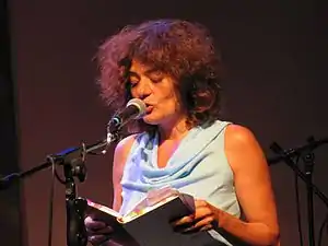 Mishori, reading her poetry. She is standing before a mic, holding a book in both hands.