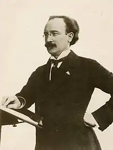 A thirtysomething man in a tuxedo and glasses clutches a violin and gazes at the camera.