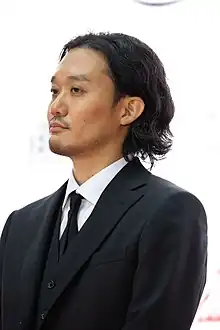 Fukunaga in three-quarter profile at press event wearing black suit and tie