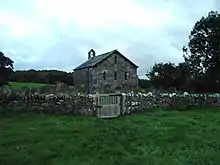 A small, simple, stone church, in front of which is a stone wall. On the far gable is a bellcote with a single bell.
