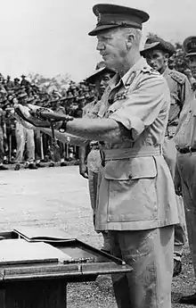 A soldier wearing a soft peaked cap presents a samurai sword over to another wearing a peaked cap with his sleeves rolled up. Another soldier in a slouch hat stands to attention nearby.