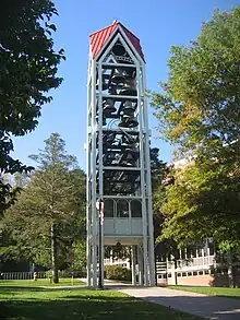 A three-dimensional vertical structure perhaps 70 feet (21 m) tall and 10 feet (3.0 m) wide by 10 feet (3.0 m) long is filled with tiers of large metal bells. The structure, standing in front of a large institutional building in a park-like setting, is supported by several metal legs. A sidewalk passes through the structure, which is open at the bottom to a height of perhaps 10 feet (3.0 m).