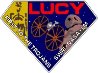 A diamond-shaped crest houses artworks of the Lucy fossil at left, the Lucy spacecraft at center, and an artist's impression of a Jupiter trojan. The word "Lucy" is written in a large, bold red font at the top right corner, while the words "First to the Trojans" and "SWRI · NASA · LM" are written in a smaller white font across the bottom edges of the diamond-shaped crest.
