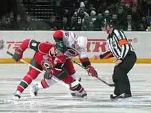 Two hokey players, one with a primarily red outfit, the other in primarily white, compete for a puck in front of a referee in an indoor stadium.