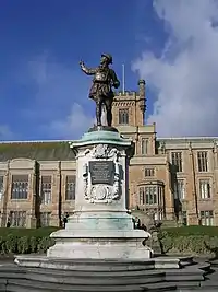 Photograph of a Gothic Revival building. The walls are light brown and the pitched roof, which has algae growing on it, is grey slate. There is a wide chimney on each side of the building. There are five large multi-segment windows on each story on the left section, and five large multi-segment windows on each story on the right section. The left and right sections have two and three stories respectively and are separated by a protruding central section, which has a total of five stories. The central section has a two-storey bay window on each side of an arched, wooden door, which is elevated. The door is above an outdoor double staircase, which has a simpler wooden door in the front. Above each bay window is a smaller window. Above the smaller windows is a narrower section with three large multi-segment windows, above which is a yet narrower section, forming a square tower and the highest storey of the building. The tower is crenellated and has one large multi-segment window, a flagpole without a flag in the centre, and a crenellated stone structure projecting from the right corner that resembles a crow's nest in that it could be used as a lookout point. In front of the building is a lawn that separates an embankment from a planted area. Steps lead down from the central part of the building to a war memorial, which is in the form of a bronze statue of a First World War uniformed British soldier looking backwards and pointing up towards the left, standing atop a white stone plinth that has a relief of the heraldic achievement of the city of Nottingham, an inscription on a plaque, and a relief of the lozenge of Dame Agnes Mellers. The war memorial is elevated with stone steps on all four sides, which separate an outer circle of shrubbery. Below the war memorial is a paved stone floor.