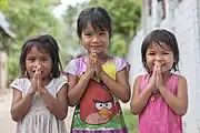 Group portrait photograph at bust length of three Lao girls joining their palms for the Thai greeting (Namaste).
