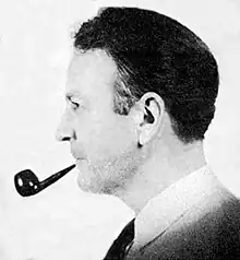 Man with slicked-back black hair facing left, smoking a pipe