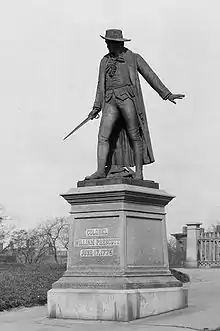 A statue of a man wearing cape and hat, and holding a sword standing to the left