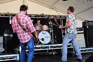 Three members of the band are seen. Two guitarists are facing away from the audience: towards the drummer who is between them at the back of the stage. The one at left wears a red, checked shirt over dark blue jeans. The drummer wears a dark cap and dark t-shirt; he is facing forward and slightly to his left. The second guitarist wears a light shirt with a montage of images over light blue jeans. Beyond the musicians is stage equipment and speakers.