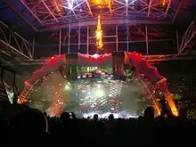 A tour stage; four large legs curve up above the stage and hold a video screen which is extended down to the band. The legs are lit up in red at the top and orange at the bottom. The video screen has multi-coloured lights flashing on it. The audience surrounds the stage on all sides.