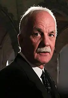 A photograph of a man with white hair and a white moustache facing the right and looking just to the left of the viewer while wearing a necktie