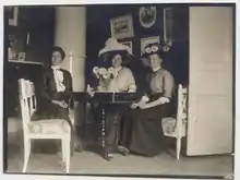 Three white women seated at a table indoors; two are wearing large hats